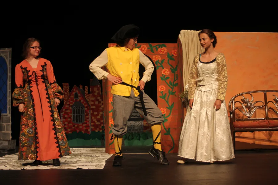 Students in costume performing Shakespeare on stage under theater lighting.